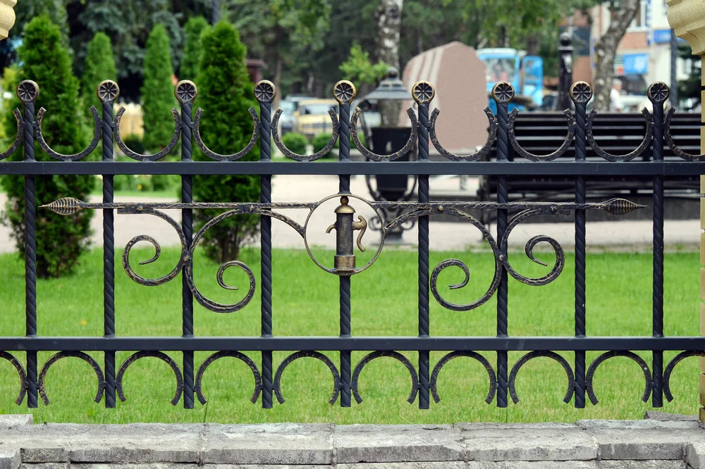 Ornate black iron fence with symmetrical swirl designs and an arrow motif. Bright green grass and trimmed trees in the background evoke a calm park setting.