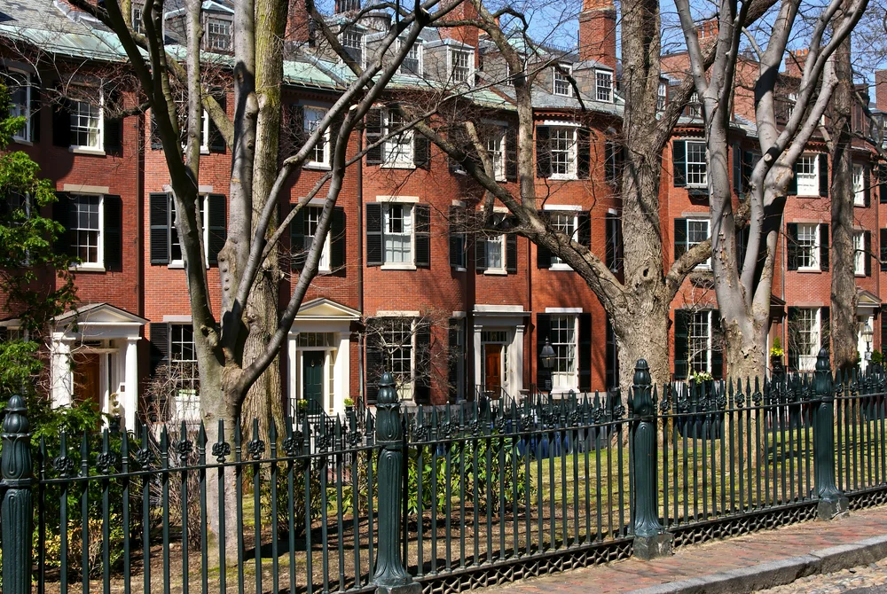 Historic red brick rowhouses with white-trimmed windows and black shutters line the street, framed by leafless trees and a decorative wrought iron fence.