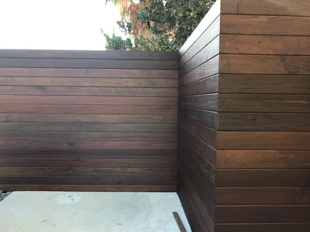 Brown wooden fence with horizontal planks forming a corner against a concrete ground. Trees are visible above, suggesting an outdoor setting.