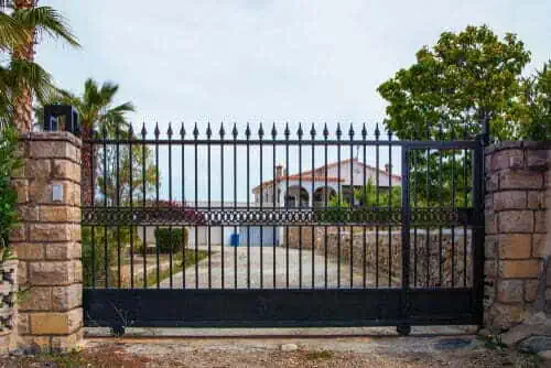 Black wrought-iron gate with pointed tops, flanked by stone pillars, leading to a white house and driveway surrounded by greenery and trees.