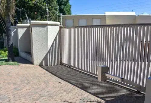 Alt text: "A tall, light-colored metal gate stands closed, casting striped shadows on a brick driveway. Behind it, a building and green trees are visible."