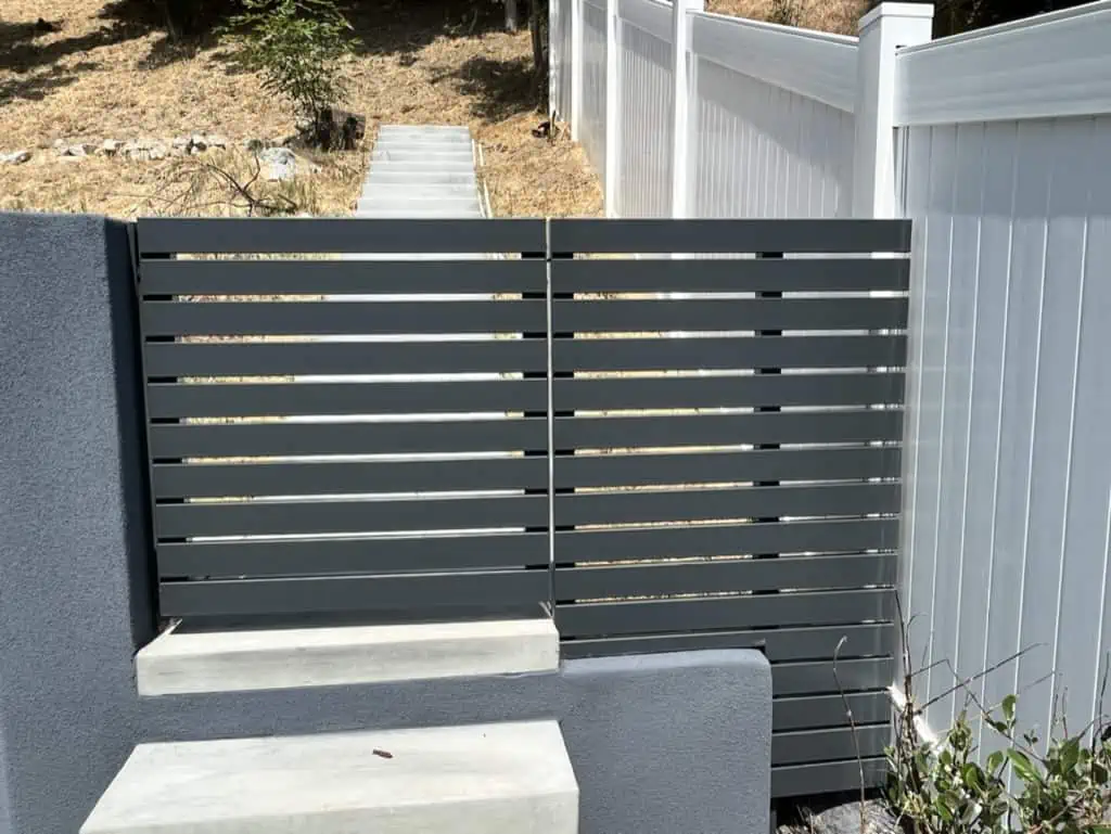 Gray horizontal metal slat gate stands between white fencing, partially open on a concrete pathway. Dry grass and trees are visible in the background.